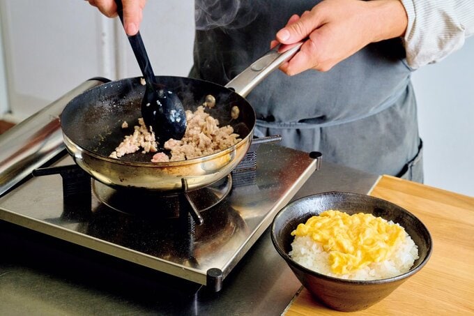 ご飯と卵が乗ったどんぶりの横で肉を炒める様子