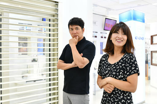 白いシャッターの横で考え込む夫とほほ笑む妻