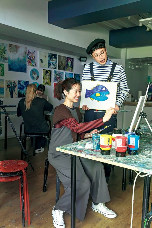 キャンバスに絵を描いている女性と、魚の画を持ちながらそれを眺める男性