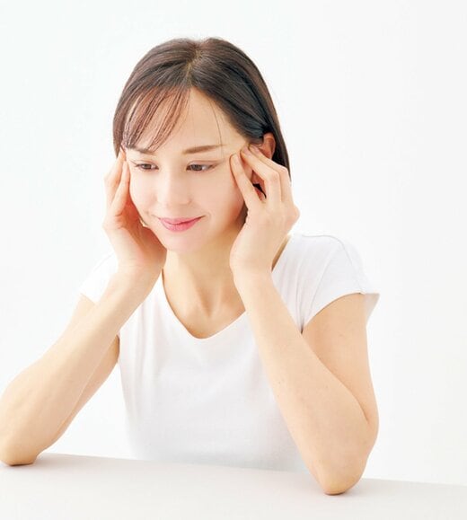 テーブルに肘をついて目尻横の頬骨の辺りを指で押し上げる女性