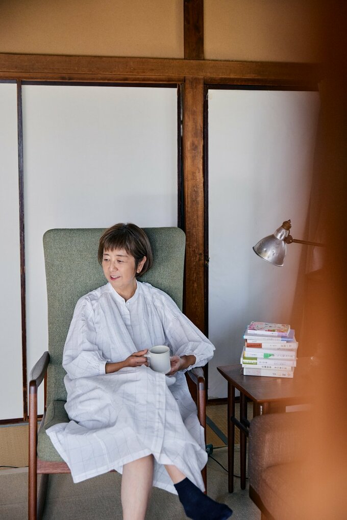 一田さんがひとり掛けソファにかけ、マグカップを持っている。傍らのテーブルに本が積んである。