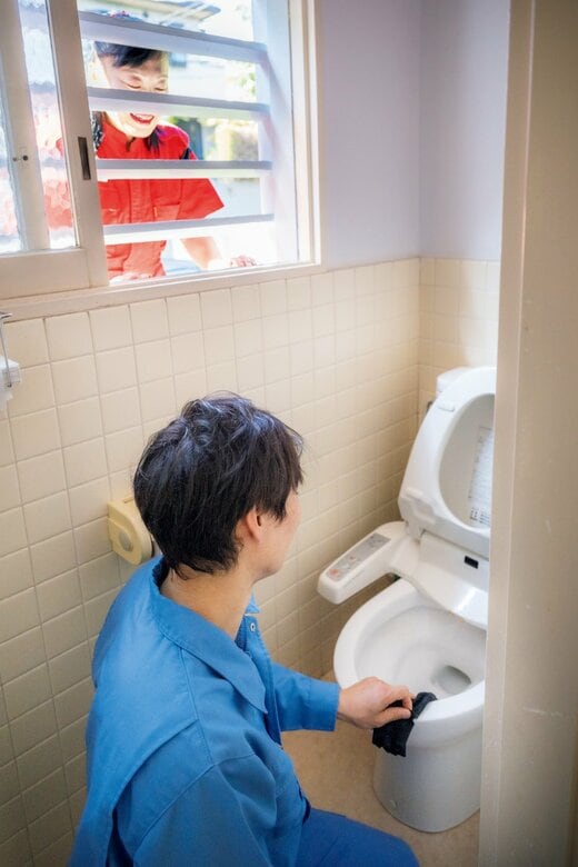 トイレ掃除をする滝沢さんと、その様子を窓から見る新津さん
