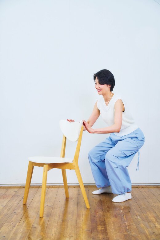 椅子の背後で背もたれに両手を置いたままヒザを曲げてスクワットする女性