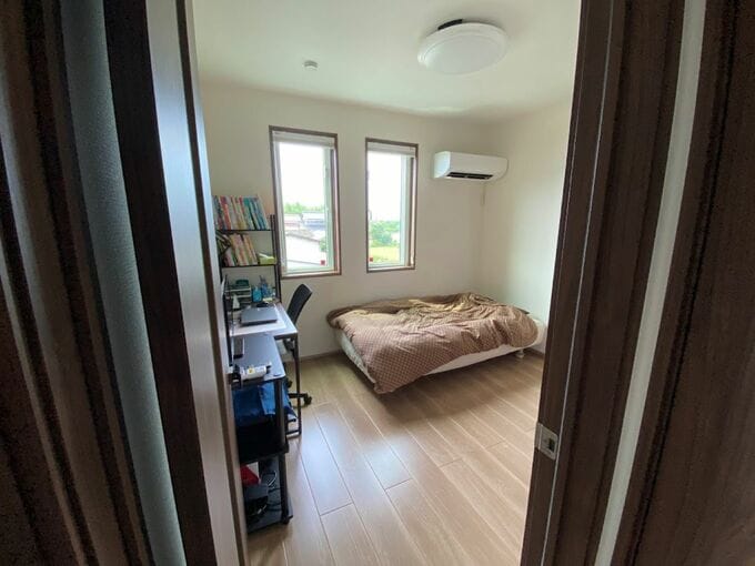 ドアから部屋を写している。右奥にベッド、左奥にデスクと本棚、奥の壁に窓がある。