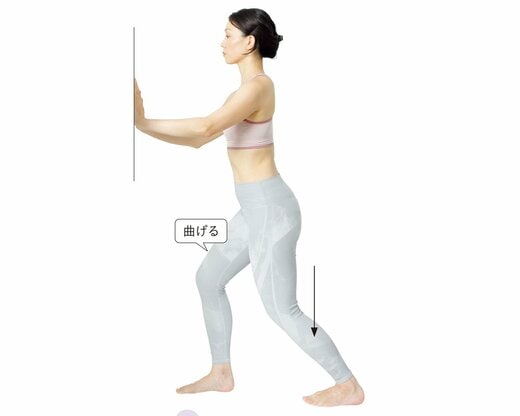 壁に手をつき、右脚を前にして脚を前後に開き、後ろ脚のひざを曲げている女性