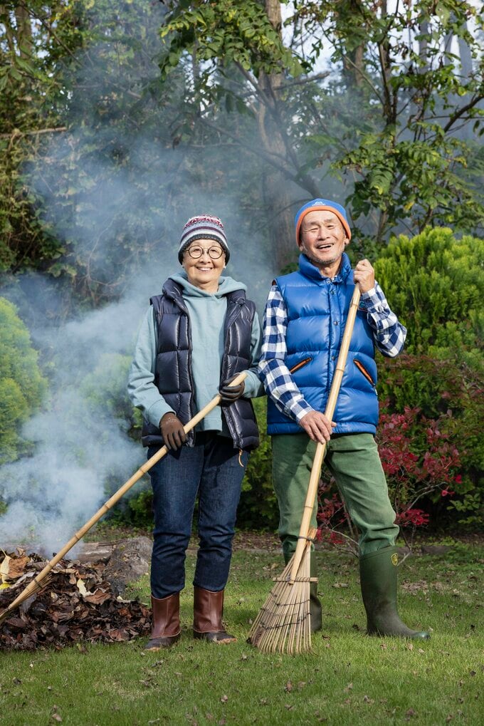 屋外で、箒を持った老夫婦が隣り合って立っている。笑顔。その左側に落ち葉があり、そこから煙が上がっている。