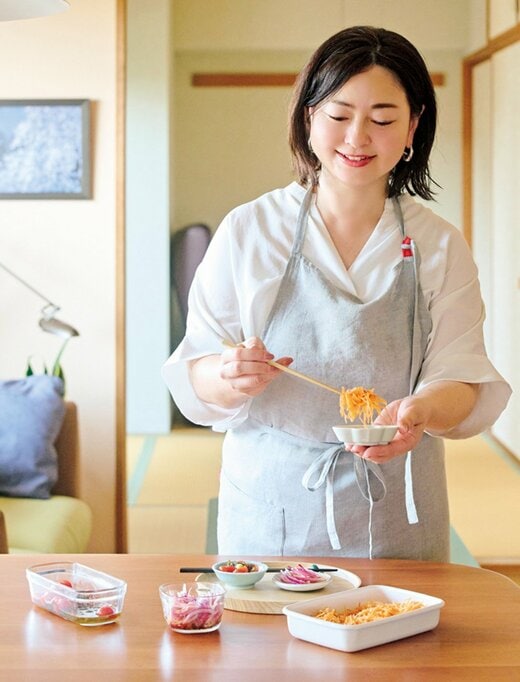 白いトップスにグレーのエプロンをかけた女性がおかずを器に入れながらほほ笑む様子